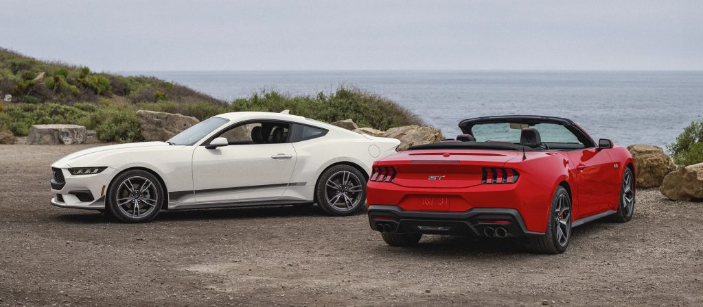 2025 Ford Mustang Mach-E lineup featuring a white coupe and a red convertible parked by the ocean, showcasing the car’s sleek design, sporty stance, and modern styling.