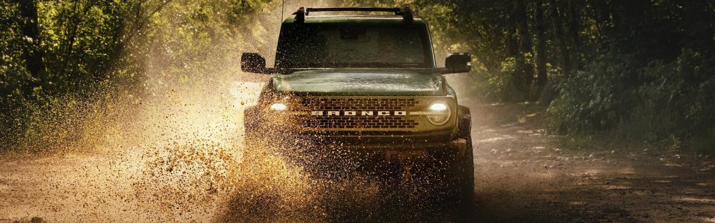A green Ford Bronco driving through a dirt trail, kicking up dust.