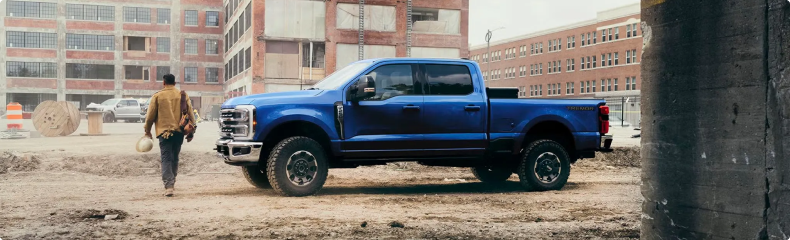 Blue Ford Super Duty Crew Cab parked on a dirt lot beside brick buildings